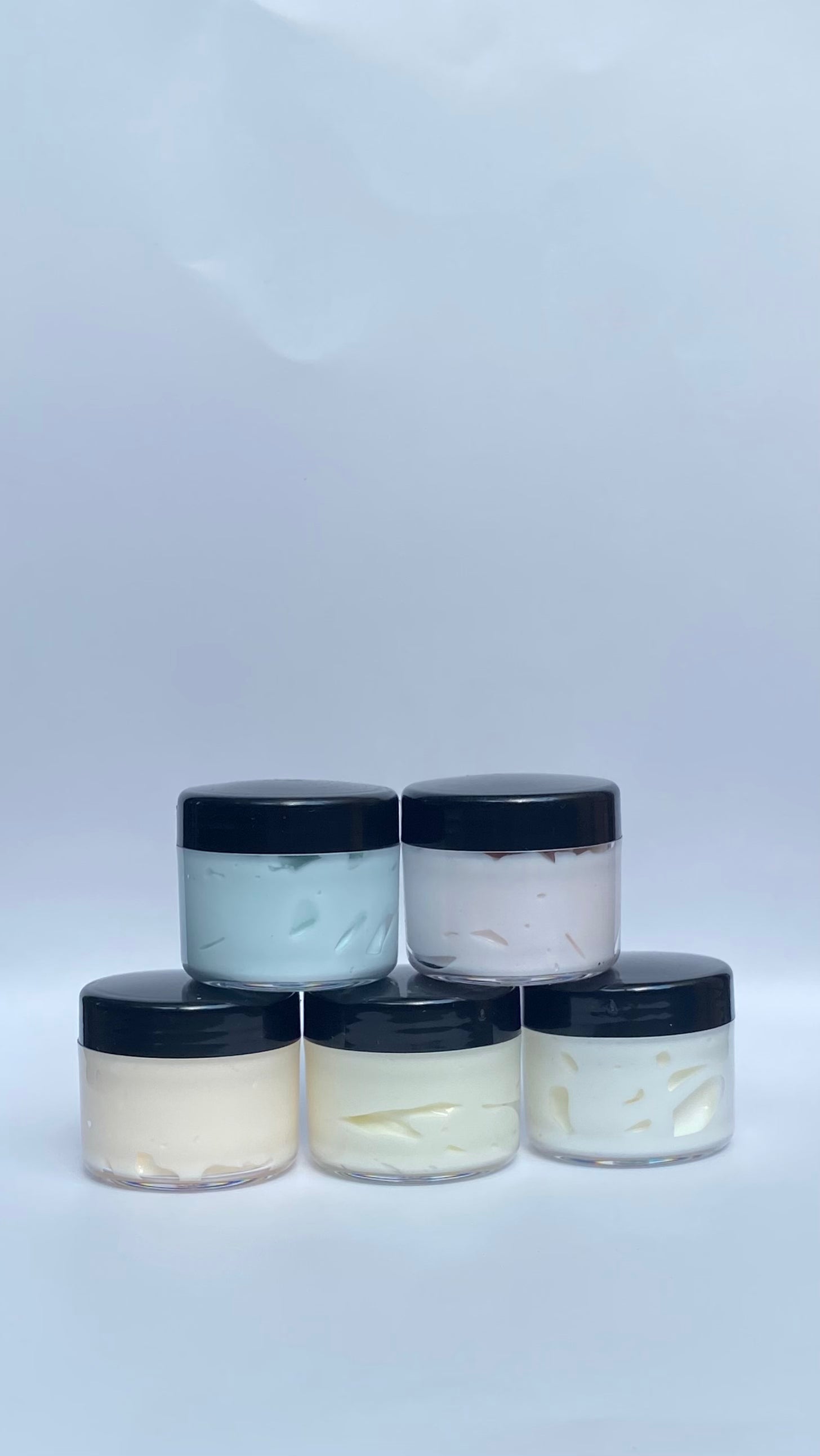 Five glass jars with black lids containing a white cream-like substance on a light gray background